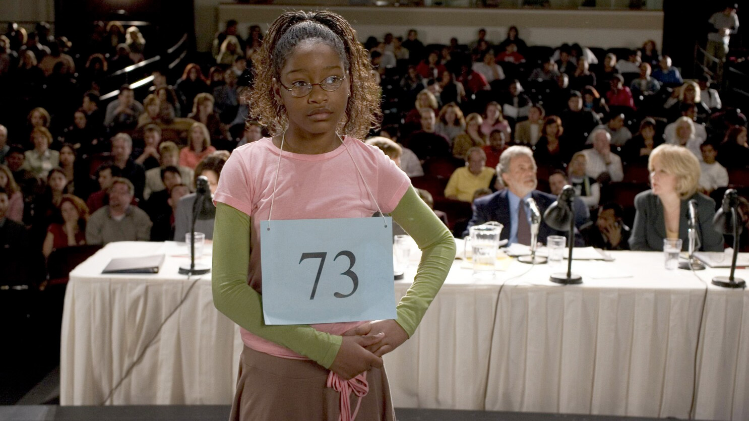 A young girl wearing a number 73 sign around her neck, indicating her competitor number for a spelling bee. She is standing in front of a panel of judges who are sitting at a table. Behind them is a sizeable audience.