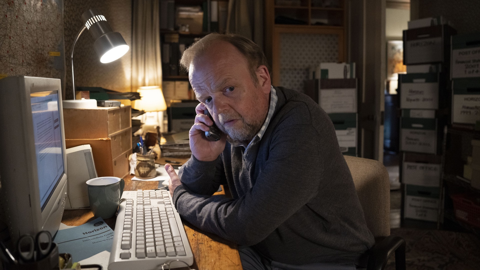 Mr. Bates sits in a dim office, on the phone, in front of a computer.