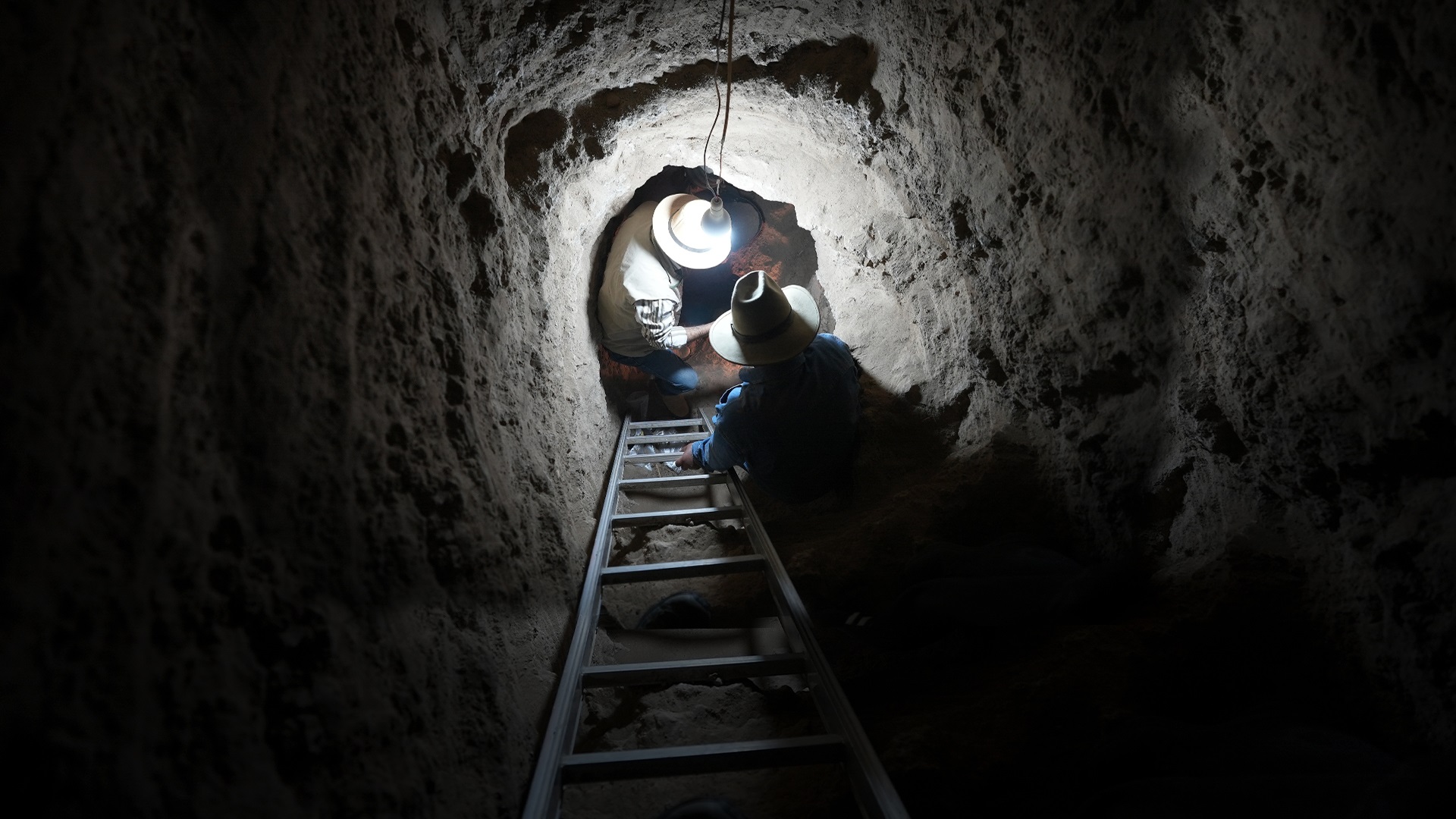 Two people in a dark Egyptian tomb shaft illuminated by a light
