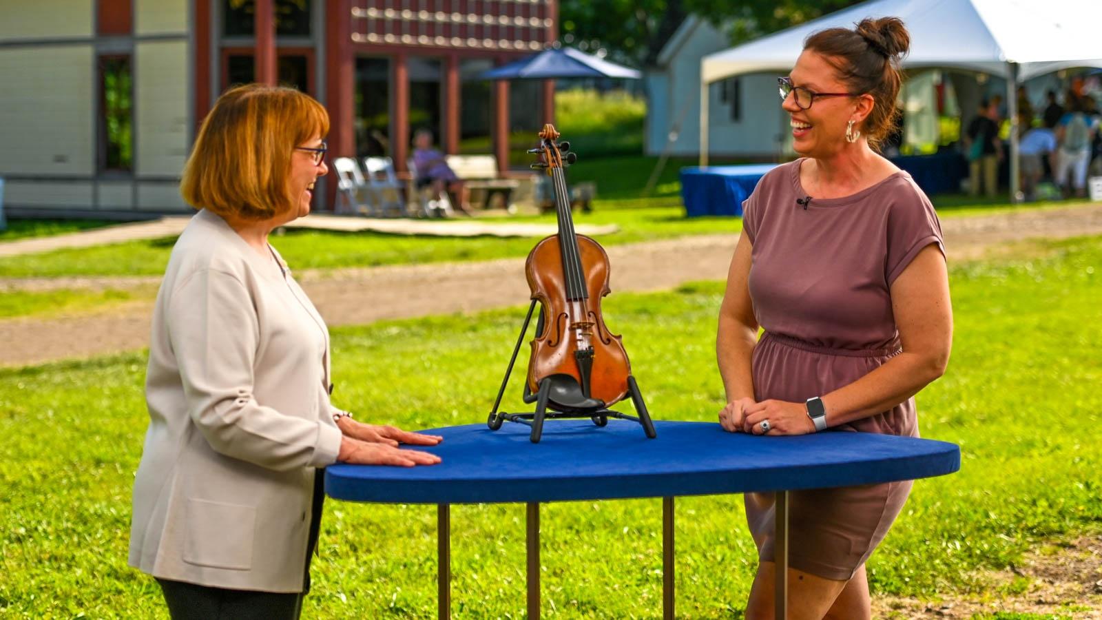 Two people stand outside on either side of a blue table where a violin is displayed