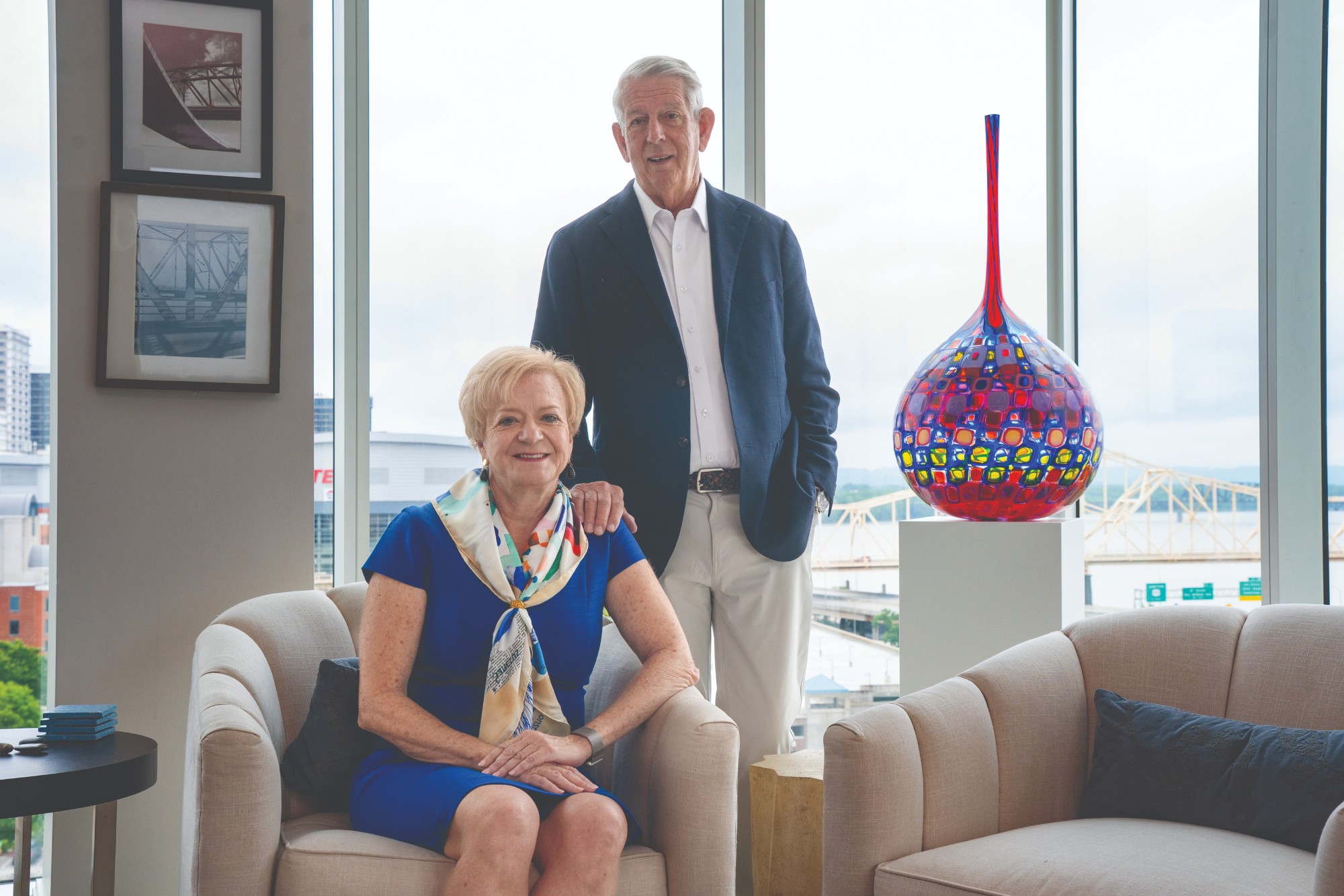 Diane Medley seated and smiling and Prewitt Lane standing behind her with his right hand on her left shoulder. Behinf them is a large multi-colored glass vessel and a bank of windows looking out over a bridge.