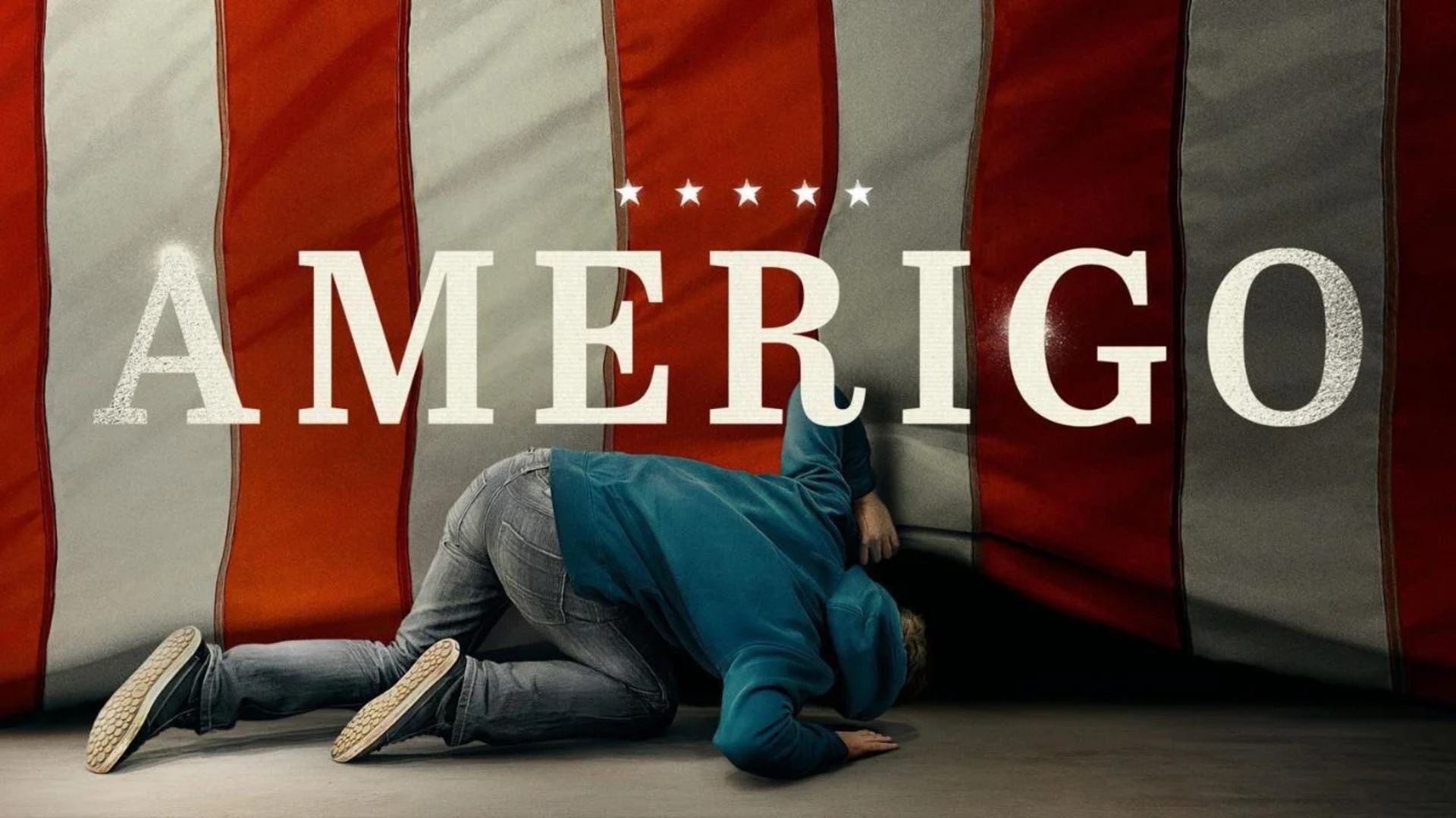 A person down on hands and knees peering under the bottom edge of a red and white striped American flag hung to the floor.