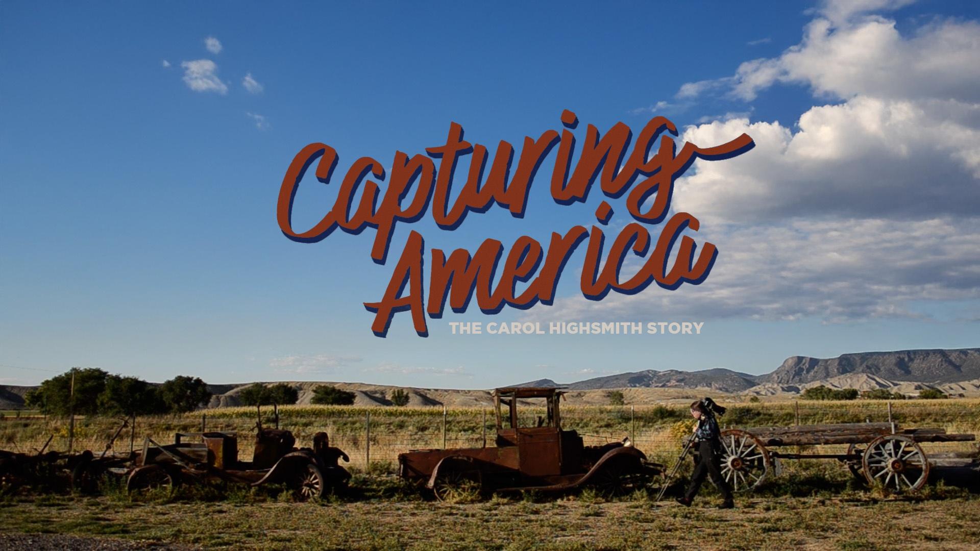 A view across a prairie with a person carrying a camera and stand walking in front of old-fashioned rusted-out cars and a wagon. In the background is a blue sky with fluffy white clouds.