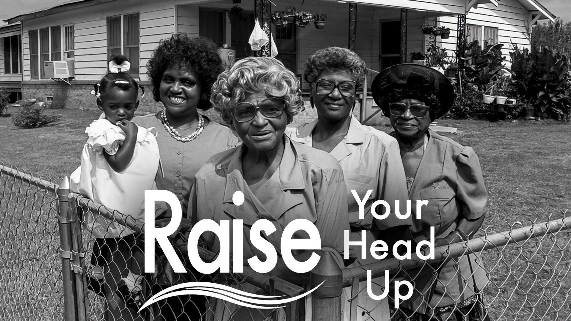 A balck-and-white photo of four Black women of varying ages stand side-by-side in a yard behind a chain-link fence, looking directly at the camera. On the far left, a younger woman holds a toddler who is resting her hand near her mouth. Behind the group stands a single-story house with a porch and surrounding trees.