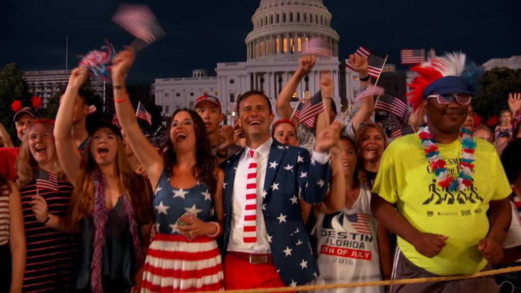 People dressed in U.S. themed apparel, including American flag-inspired clothing and festive accessories like hats and leis. They are smiling and waving small U.S. flags. In the background in the U.S. capitol dome.