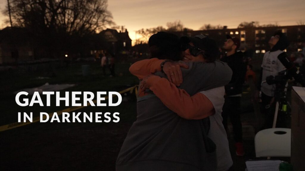 People hugging in dim outdoor light during an eclipse. The program title treatment is superimposed over the image.
