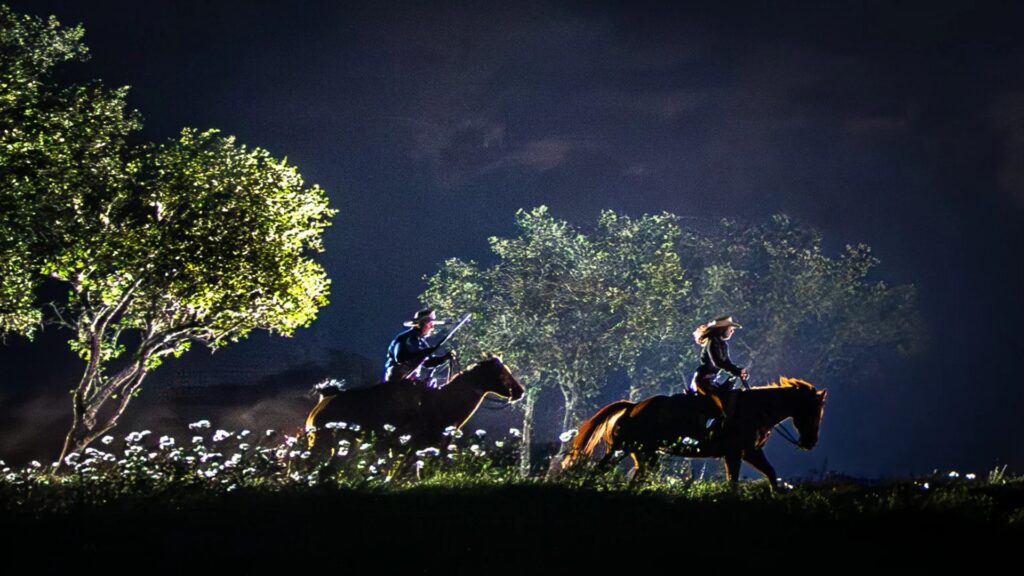 People riding horses through trees under a dimly lit sky during an eclipse.
