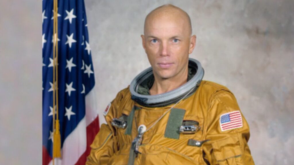 Astronaut Dr. Story Musgrave in an orange spacesuit with a US flag in the background