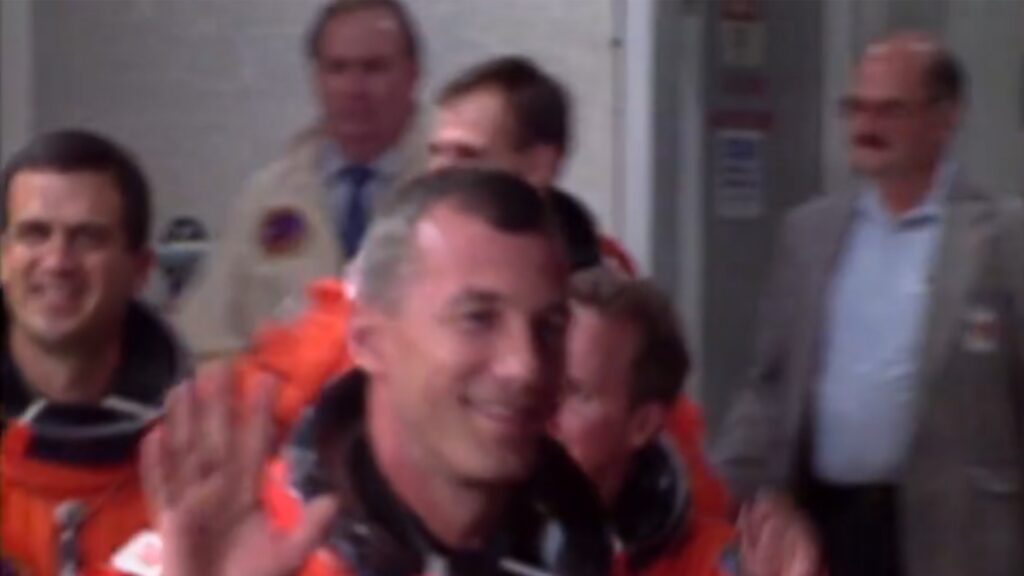Astronaut Terry Willcutt waving in an orange spacesuit with other astronaurts and people in the background