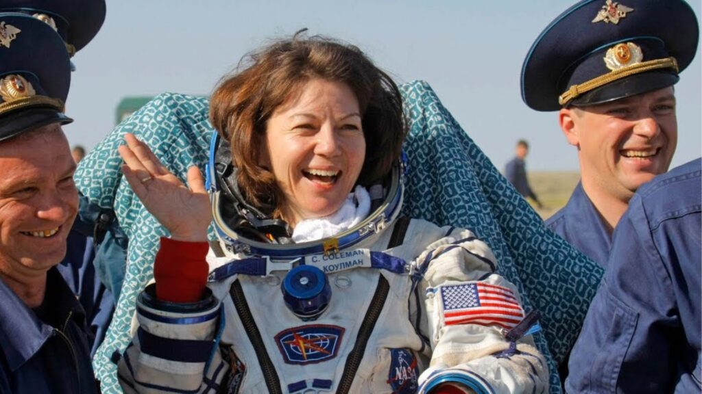 Astronaut Cady Coleman in a space suit waving and smiling with two men in military uniforms on either side of her