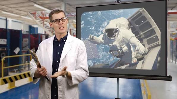 A man wearing a white lab coat standing in front of a large monitor with an astronaut on it