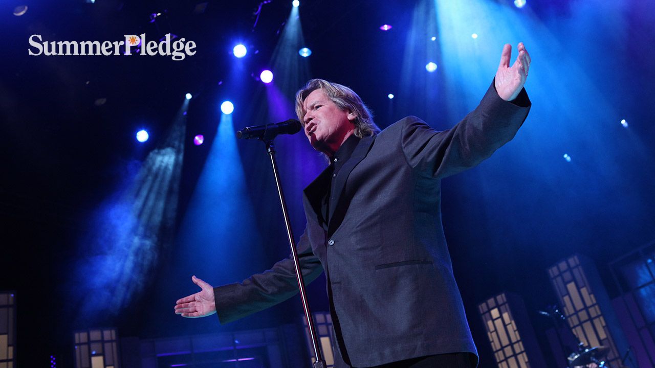 Man singing on stage with arms outstretched, blue light background