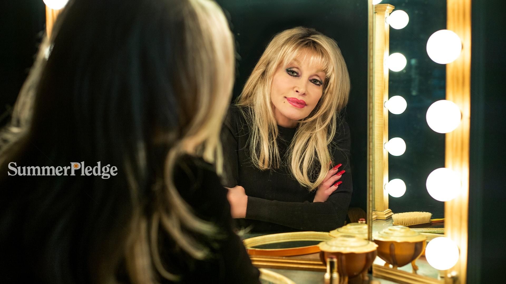 Dolly Parton sitting at a makeup table looking in a mirror rimmed with small light bulbs.