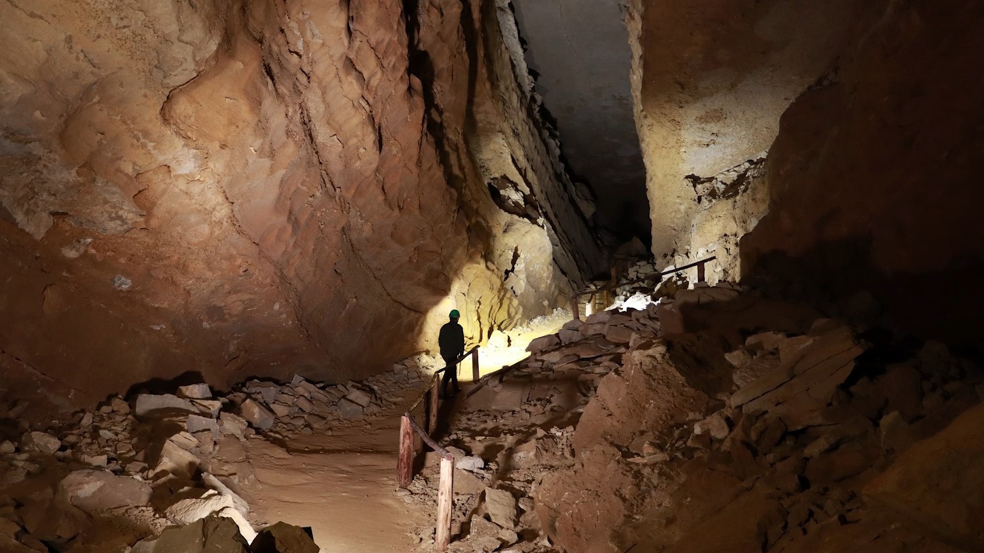 A person descends down a path alongside a handrail into Mammoth Cave. In the background are rock walls and large rocks.