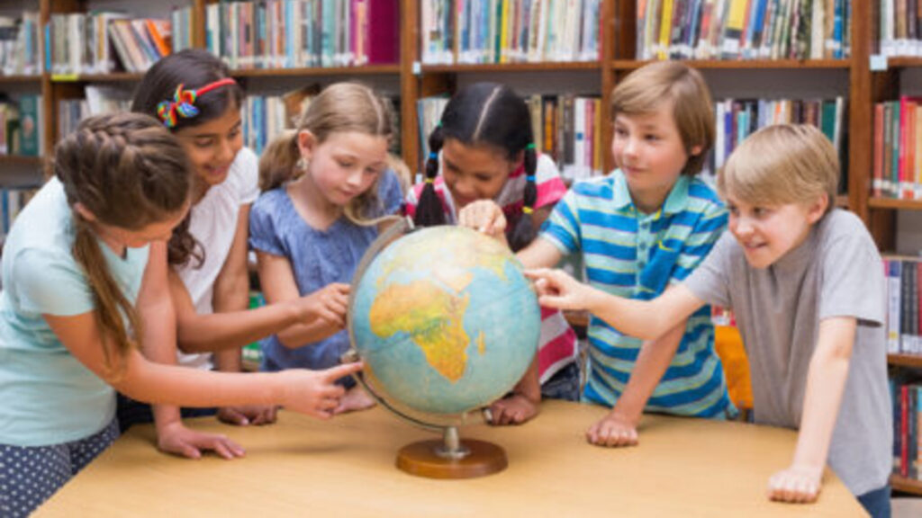 Six young children looking at and pointing to various places on a globe which is sitting on a table in a library. Behind them are tall wooden bookshelves with books that have white labels at the bottoms of their spines.