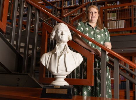 Rae Ann Sauer in a green patterned dress standing on a stairwell with a white bust of a man in front of her and books on shelves behind her.