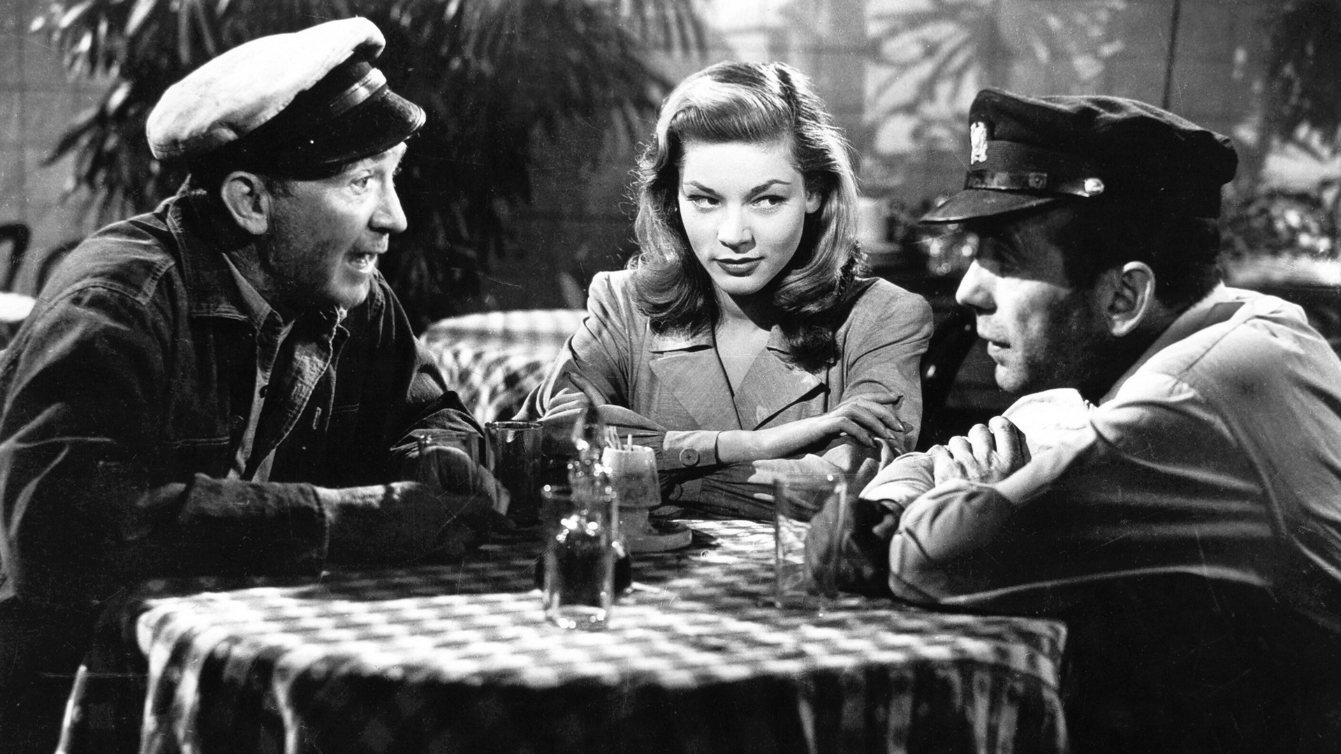 Black and white still from film with Brennan, Bacall, and Bogart around a table with a checkered tablecloth