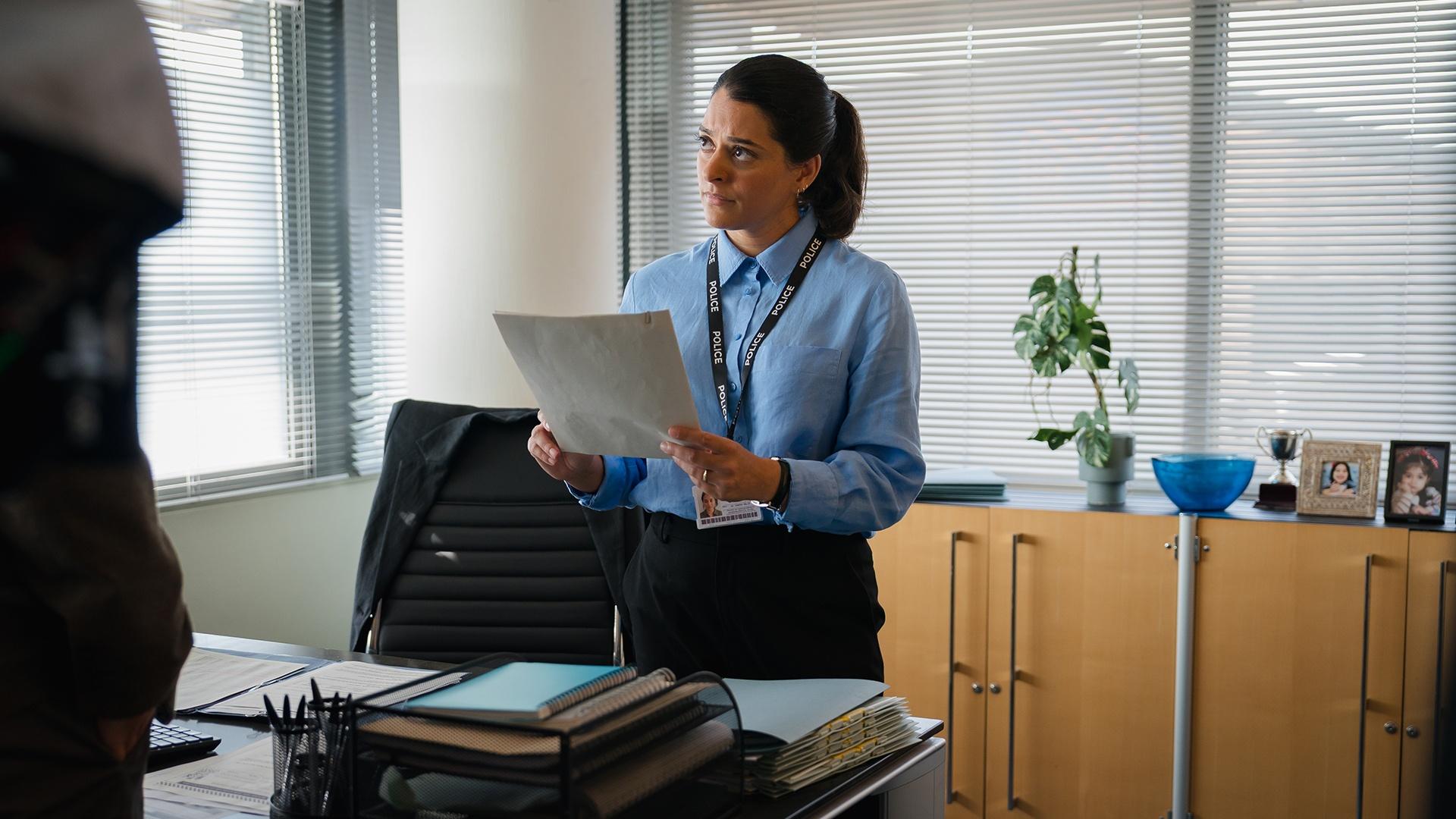 DI Malik behind a desk holding a file