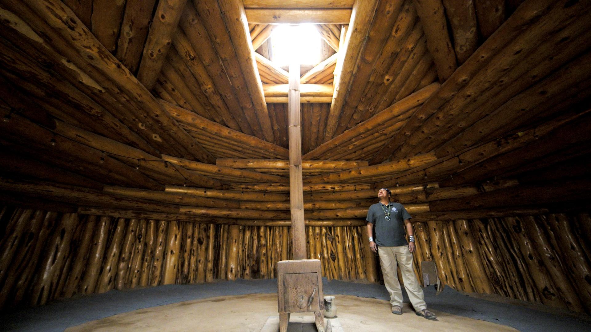 Native American stands in a wooden building with a rounded ceiling with an opening in the middle