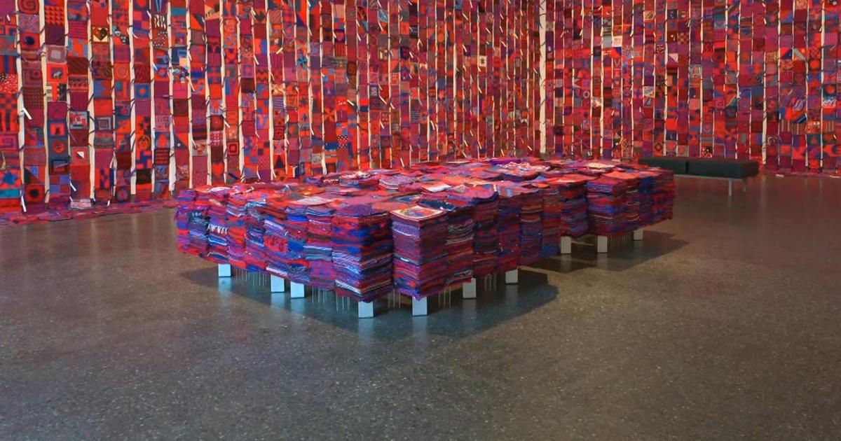 An exhibit of red and blue quilt squares covering two walls with pallets of more squares stacked on the floor in front