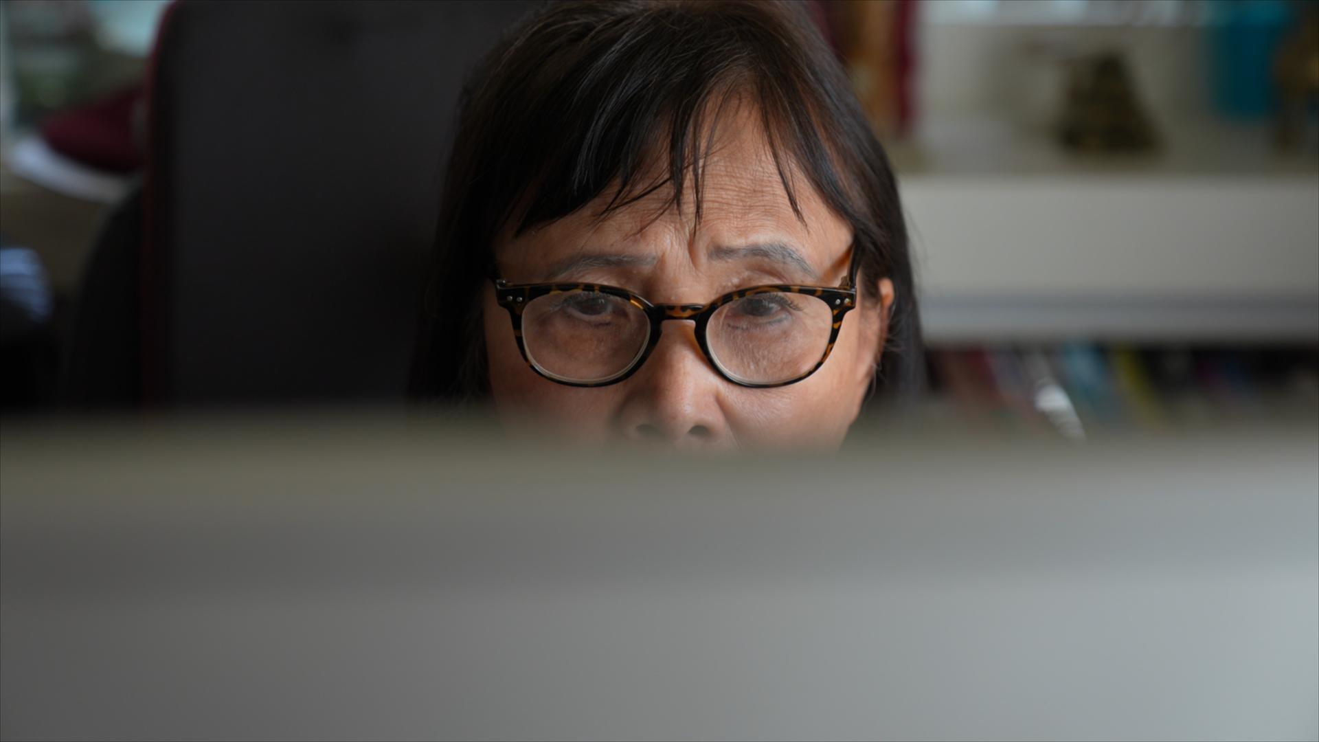 An older person wearing glasses looks at a computer monitor, which is covering the lower half of their face