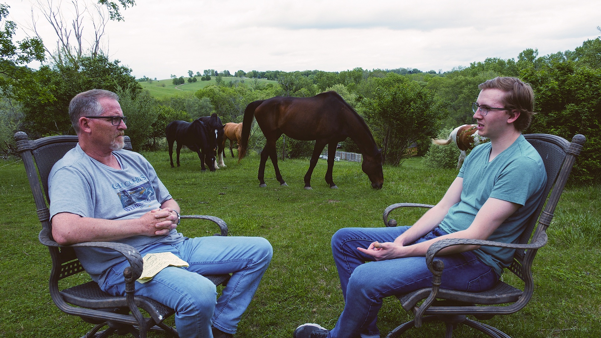 Filmmaker and his father sit across from each other outside, farm animals in the background