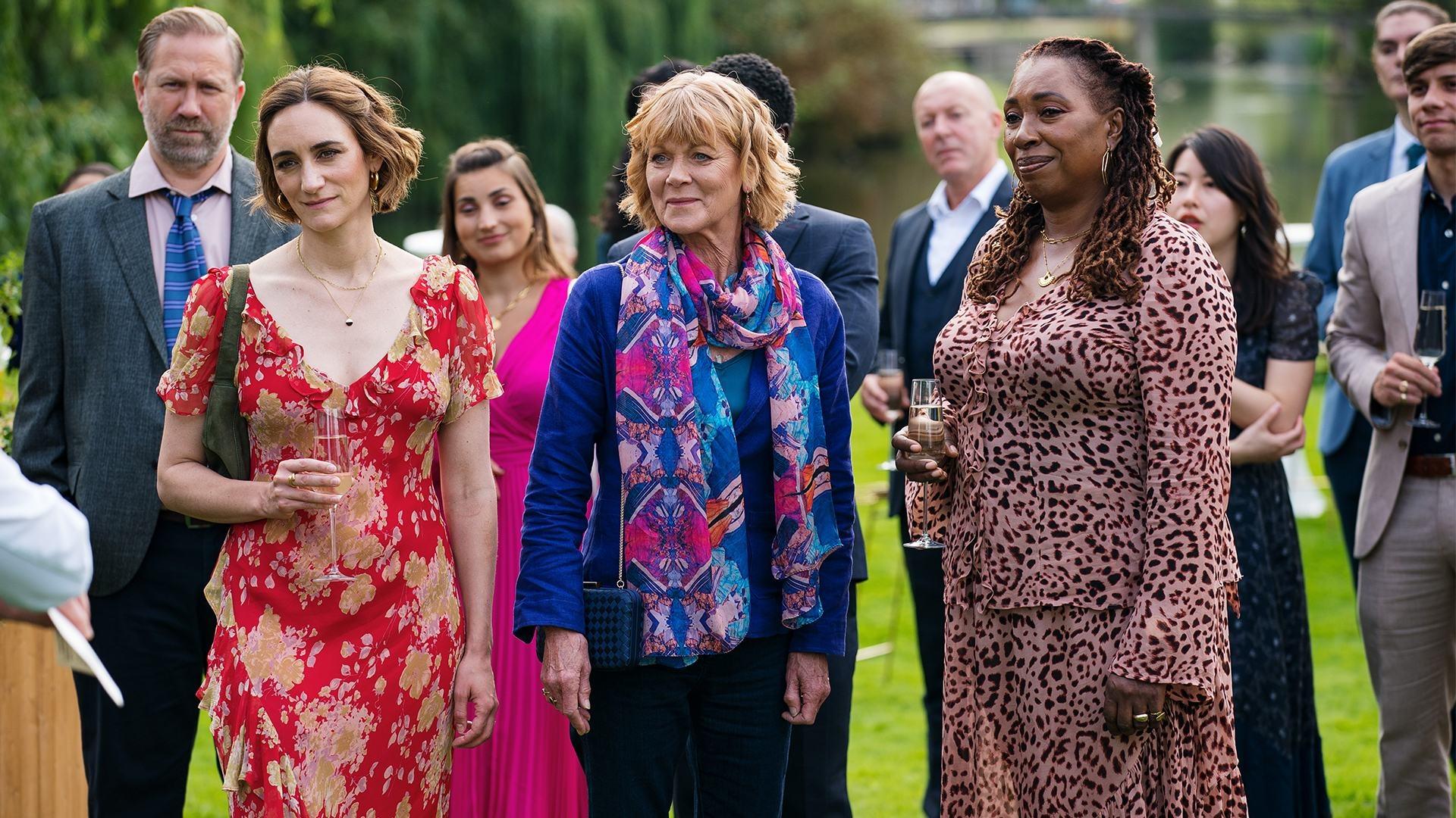 Becks, Judith, and Suzie stand with other people outside in dressy clothes, holding champagne flutes