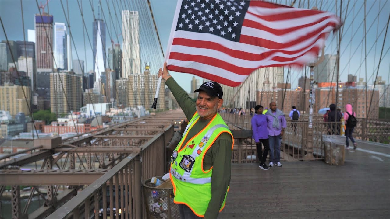 A person in the Walk with Frank event holding a large American flag while walking across the Brooklyn Bridge in New York City