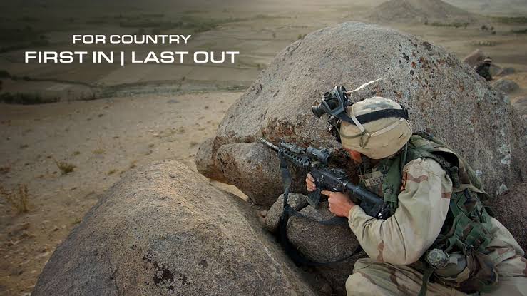 A U.S. Marine in desert camouflage crouched behind a large rock, aiming a rifle in a rugged, arid landscape.
