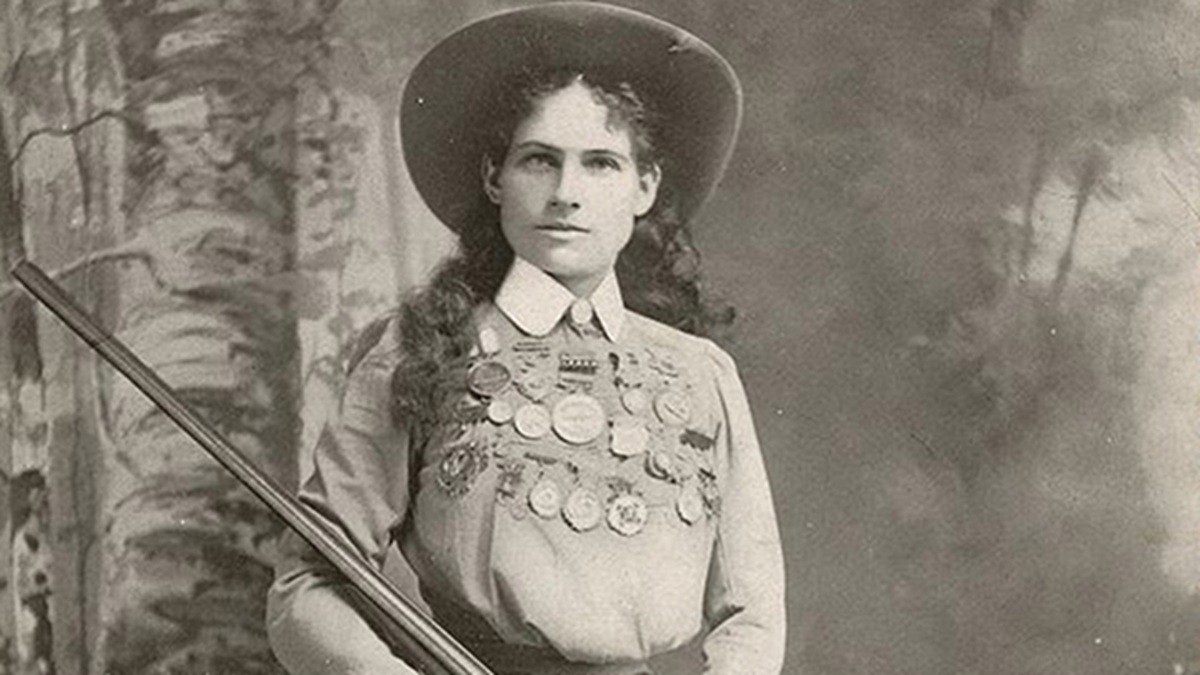 black and white photo of Annie Oakley with medals on her chest, wearing a hat, and holding a rifle