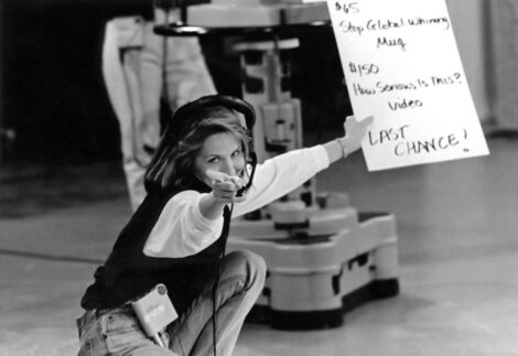 Shae Hopkins crouching next to a TV camera and holding a cue card in her left hand while pointing with her right hand at a photographer who is taking her picture.
