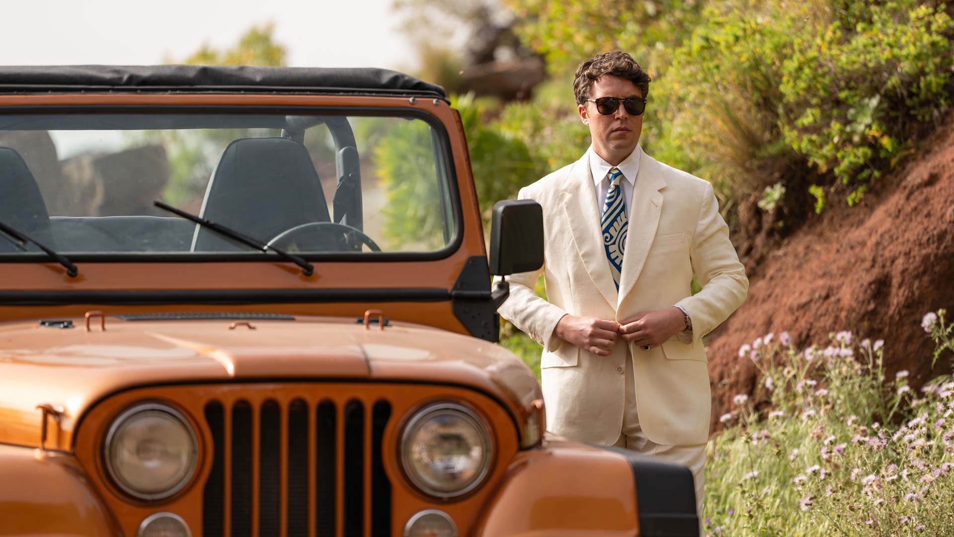 Logan buttoning a white suit jacket, standing next to an orange jeep