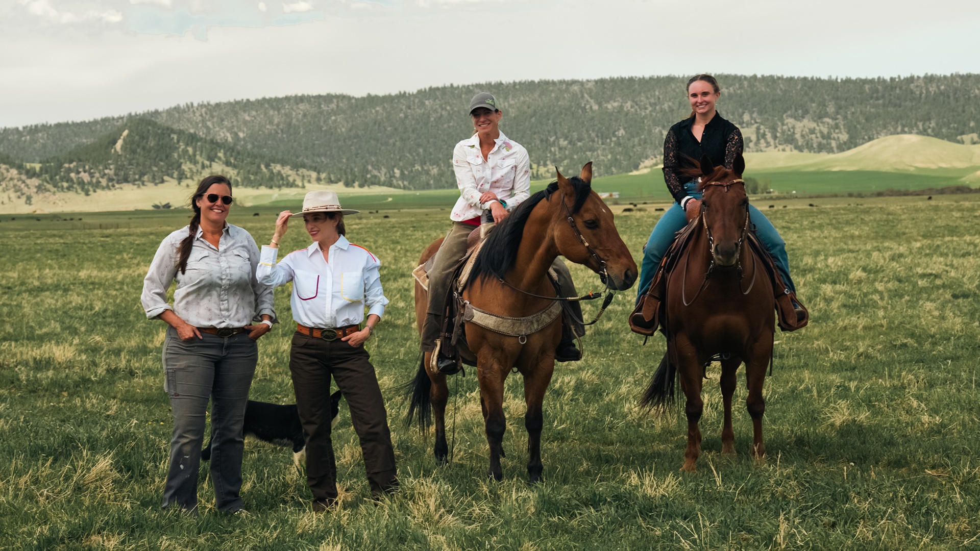 Pati and another person stand next to two people on horseback on grassland with mountains in the background