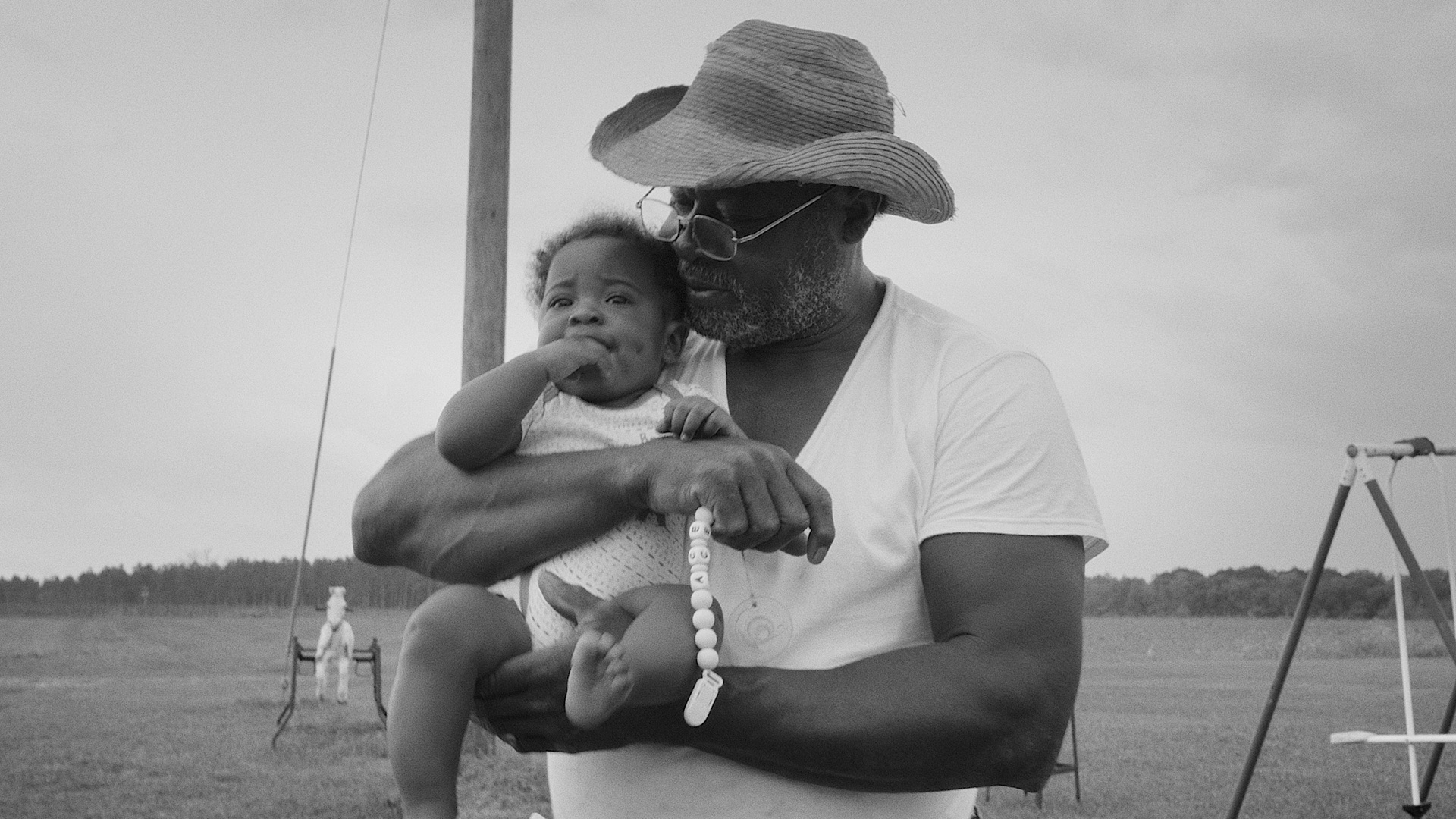 An older man in a wide brimmed hat holds a baby, a field in the background